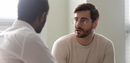 Man consulting with practitioner, African psychologist holding clipboard with card sitting in front of patient listens to his mental health complaints.  Job interview process applicant and HR manager concept