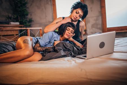 Two girls, young lesbian couple relaxing together at home in bed, using laptop.